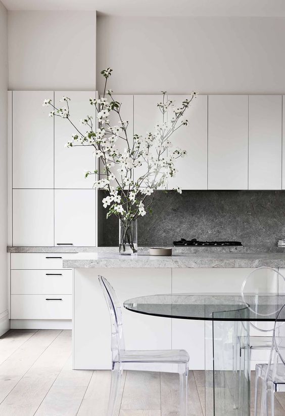 Beyaz renkteki zarif minimalist mutfak gri tas duvar ve tezgahlarla cam masa ve akrilik sandalyelerle tasarlanmistir