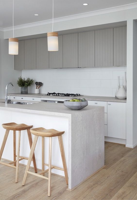 Sik ve minimalist bir mutfak beyaz dolaplar ve gri ust dolaplar beyaz fayans arka zemin mutfak adasinda su perdesi tezgah ve sarkit lambalar bulunuyor